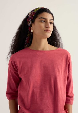 Donna con lunghi capelli scuri, che indossa una maglietta rossa e un foulard floreale in testa, guarda verso destra con un'espressione neutra su uno sfondo semplice.