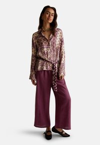 Frau steht in einem cremefarbenen und lila blumengemusterten Shirt mit gebundenem Gürtel, weiten lila Hosen, schwarzen Netzschuhen und goldener Schmuck.