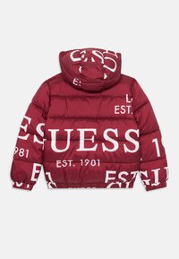 Burgundy puffer-jacka med huva, prydd med stora vita texter "GUESS" och "EST. 1981" tryckta över hela, med quiltad design och elastiska ärmslut.