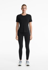 Egy nő áll szemben, fitted fekete rövidujjú pólóban, fekete leggingsben és világosszürke futócipőben, fehér háttér előtt.