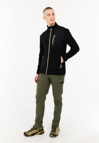 Uomo in piedi che guarda a sinistra, indossando una giacca nera con zip, pantaloni verde oliva con cerniera sulla tasca e scarpe da trekking beige su sfondo bianco.