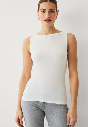 Vrouw in een mouwloos wit shirt en lichtgrijze spijkerbroek staat voor een effen lichte achtergrond en kijkt recht vooruit.