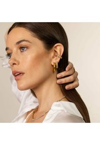 Goldene Creolen und funkelnde Kristall-Ohrenkappen schmücken das Ohr eines Modells und ergänzen ihre glänzende weiße Bluse sowie ihre glatte Frisur.