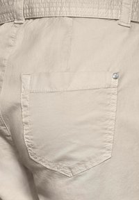 Tissu beige clair avec une texture lisse, présentant une poche arrière de forme pointe et un accent en métal. Détails de couture présents.