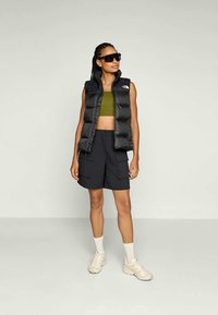 Svart pufferväst, olivgrön crop top, mörka cargoshorts och ljusa sneakers. Modell som bär solglasögon; neutral bakgrund.