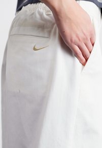 Bele hlače s elastiko v pasu, ki imajo majhen zlat Nike logotip na zadnjem žepu in stranski žep na desni strani.
