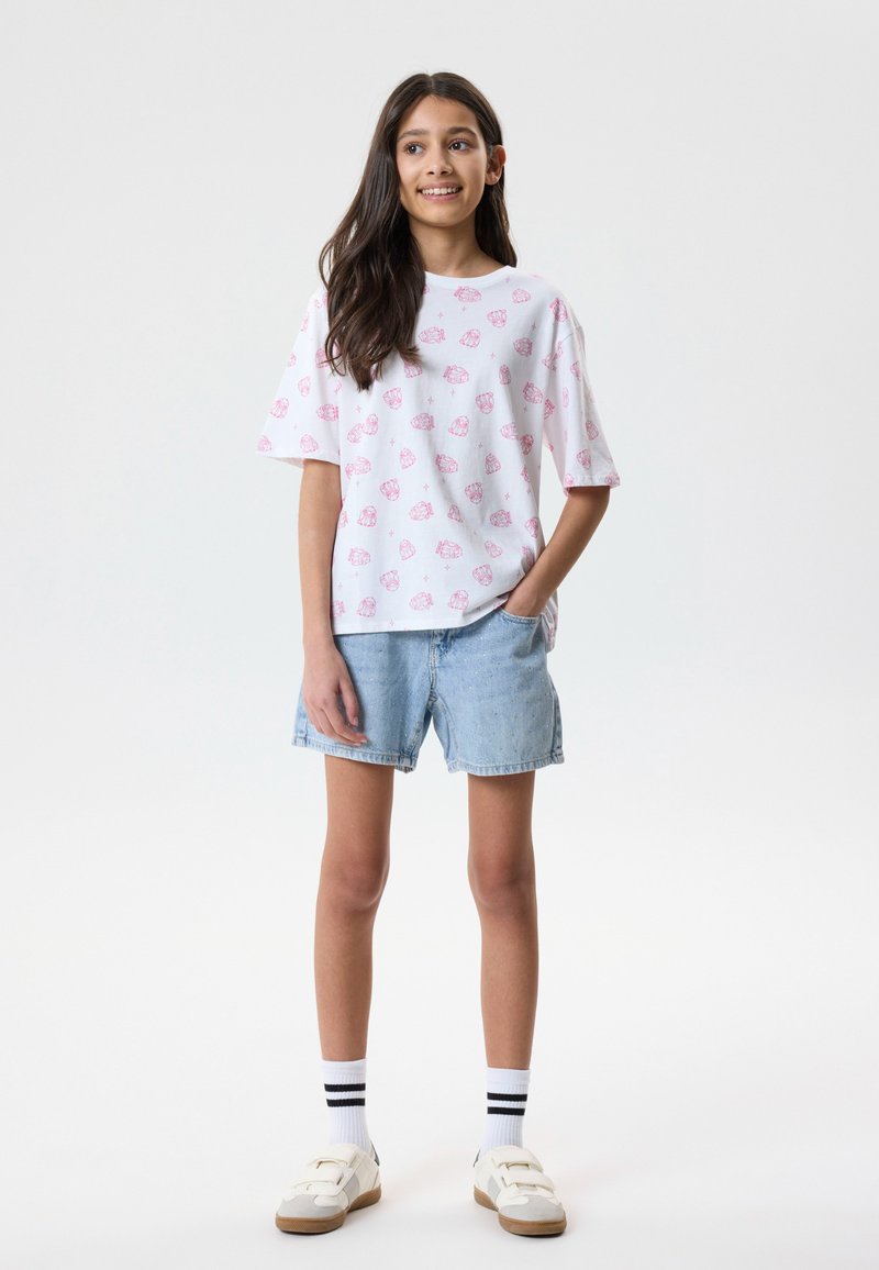 Niña con cabello largo y oscuro, vistiendo una camiseta blanca con patrones rosas, pantalones cortos de mezclilla claros, calcetas blancas con rayas negras y zapatillas deportivas blancas.