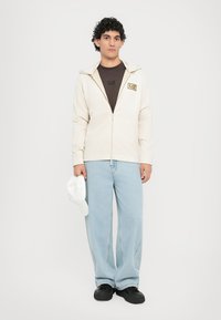 Felpa con zip color beige chiaro con cappuccio, T-shirt nera sotto, jeans a gamba larga blu chiaro e scarpe nere, che tiene in mano un cappellino bianco.