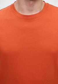 Personne portant un t-shirt orange uni à col rond, ne montrant que le cou et le haut de la poitrine.