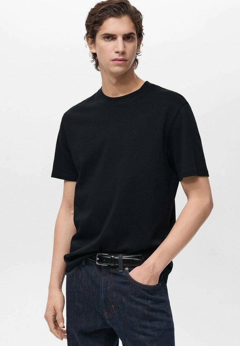 Tânăr cu păr brunet, îmbrăcat cu un tricou simplu negru cu mânecă scurtă, blugi închiși la culoare și o curea neagră, cu o mână în buzunar.