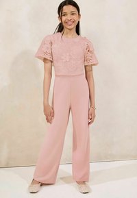 Fille souriante, portant un haut en dentelle rose à manches courtes avec des motifs floraux et un pantalon large assorti, se tenant devant un fond beige.