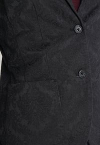 Blazer noir avec un motif jacquard floral subtil, deux boutons noirs et une poche sur le côté gauche, porté sur un vêtement rouge foncé.