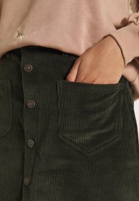 Gonna in corduroy verde con righe verticali, bottoni frontali e una tasca visibile. Abbinata a una maglia a maniche lunghe rosa chiaro.