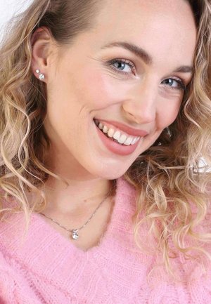 Silver necklace with a clear pendant, pink sweater with textured knit, and small stud earrings reflecting light. Curly hair framing the face.