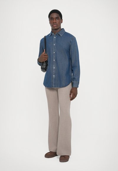 Chemise en denim bleu foncé �à boutons, associée à un pantalon beige clair évasé et des chaussures brunes à lacets. Le modèle tient un petit sac noir.