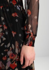 Gros plan sur une manche et une jupe de robe noire transparente avec des motifs floraux rouges et beiges et des pois blancs, avec des poignets et une taille élastiqués smockés.