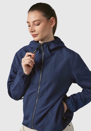 Veste légère bleu marine avec capuche, dotée d'une fermeture éclair frontale, de poignets élastiques et de poches latérales. Le matériau a une texture lisse et mate.