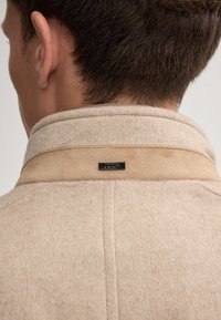 Beige wollen-blend jas met een gestructureerde afwerking, met een contrasterende beige kraag en een zwart logo label aan de achterkant.