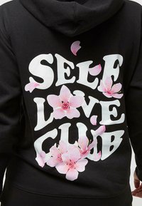 Sweat à capuche noir avec le texte blanc "SELF LOVE CLUB" et des graphiques de fleurs de cerisier roses. Tissu lisse, coupe décontractée et design à capuche.
