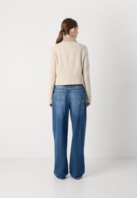 Beige strukturierter Blazer mit Kragen, kombiniert mit weit geschnittenen blauen Denim-Jeans. Sichtbare Gesäßtaschen an den Jeans, glattes Material.