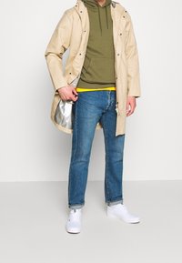 Impermeabile beige sopra una felpa verde oliva, abbinato a jeans blu e sneakers bianche. L'impermeabile ha un rivestimento argentato e hardware visibile.