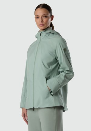 Donna con giacca con cappuccio verde chiaro e pantaloni abbinati, in piedi di profilo con una mano nella tasca della giacca, su sfondo neutro.