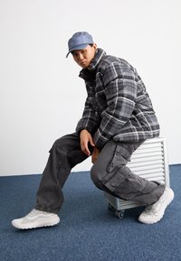 Chaqueta de plumas en cuadros negros y grises, pantalones cargo grises y zapatillas blancas. Modelo sentado sobre un contenedor de almacenamiento plateado, con fondo de moqueta azul.