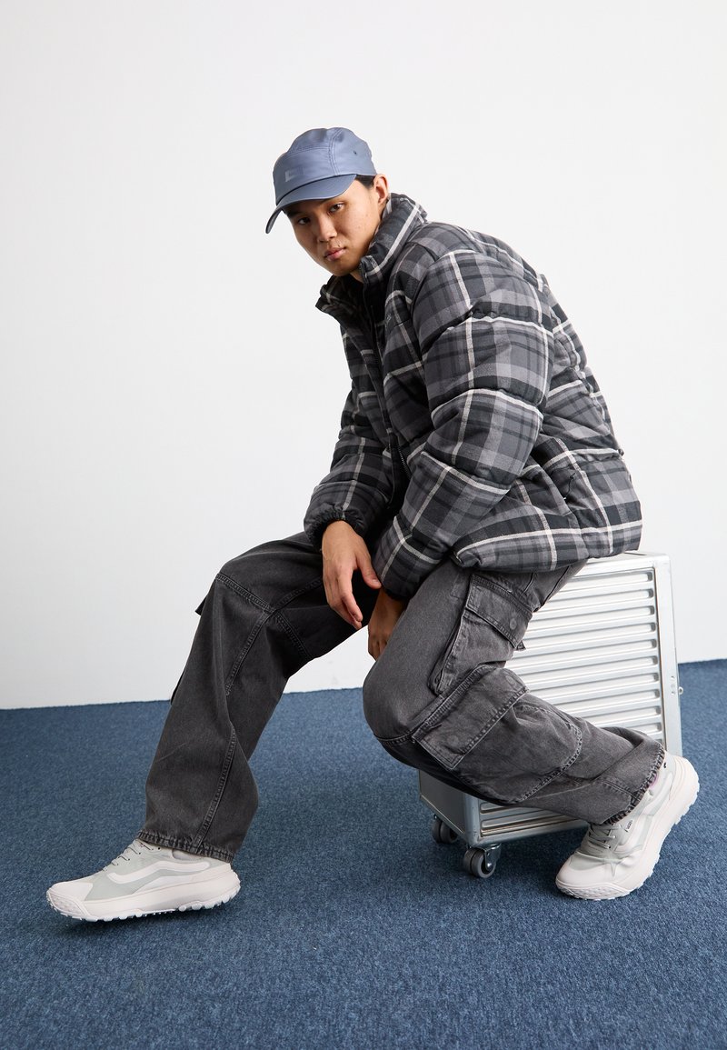 Chaqueta de plumas en cuadros negros y grises, pantalones cargo grises y zapatillas blancas. Modelo sentado sobre un contenedor de almacenamiento plateado, con fondo de moqueta azul.