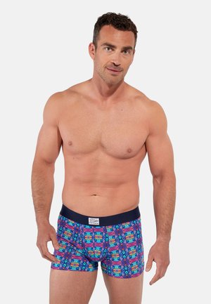 Mężczyzna bez koszulki, o krótkich ciemnych włosach, nosi kolorowe bokserki z wzorem i ciemnym paskiem w pasie na jednolitym, jasnym tle.