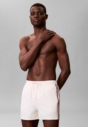 Athletischer Mann mit dunkler Haut, der hellfarbene Badeshorts mit Seitenstreifen trägt und mit einer Hand auf der Schulter vor einem schlichten grauen Hintergrund posiert.