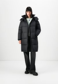 Woman wearing a long black puffer coat, black pants, black chunky shoes, and a light-colored sweatshirt, standing against a white background.