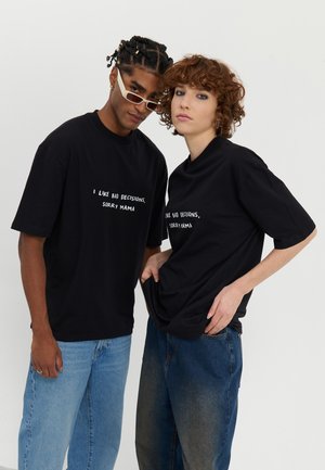 Two individuals wear black cotton t-shirts with white text: "I LIKE BAD DECISIONS, SORRY MAMA." They pair with light and dark denim jeans.