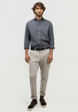 Hombre vestido con camisa abotonada gris oscuro, pantalones beige con puños enrollados, cinturón negro y zapatillas negras con suelas blancas, de pie sobre un fondo liso.
