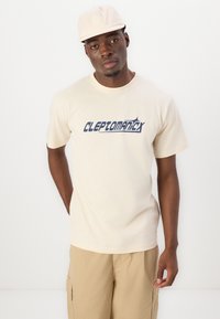 Beige cotton t-shirt with blue "CLEPTOMANIX" graphic print. Short sleeves and a crew neckline, paired with tan cargo shorts.