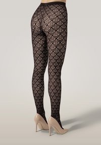 Ben klädda i svarta mönstrade strumpbyxor och beige högklackade skor mot en neutral bakgrund.