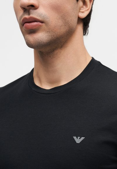 Κοντινό πλάνο άνδρα που φοράει μαύρο μπλουζάκι με στρογγυλή λαιμόκοψη και μικρό ανοιχτό γκρι λογότυπο στο αριστερό στήθος.