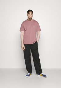T-shirt à manches courtes couleur rose poussiéreux avec une poche, associée à un pantalon noir et des baskets bleues avec des rayures jaunes. Design simple, en tissu coton.
