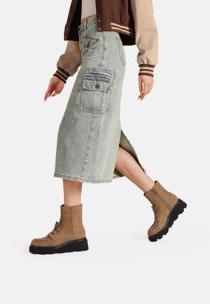 Gonna maxi in denim azzurro chiaro con spacco laterale e tasche applicate, abbinata a stivali alla caviglia in suede color tan con suole nere a piattaforma.