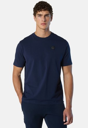 Uomo con capelli ricci che indossa una maglietta a maniche corte blu navy con girocollo e pantaloni abbinati, in piedi con le mani rilassate.