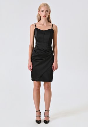 Robe en satin noir avec un corsage ajusté, des bretelles spaghetti et une jupe drapée asymétrique. Assortie à des talons noirs en vernis et des brides de cheville.