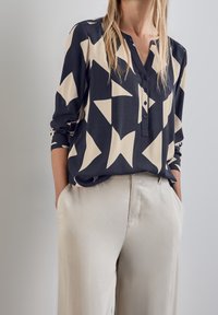 Blusa nera e beige con motivi ad astratto triangolare, maniche lunghe, patta dei bottoni e vestibilità rilassata, abbinata a pantaloni chiari.