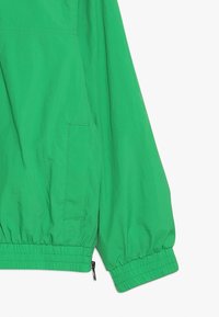 Zielona, lekka kurtka z elastycznymi mankietami i dolnym wykończeniem, z boczną kieszenią oraz gładką, lekko błyszczącą teksturą.
