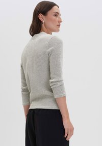 Donna con capelli castani lunghi fino alle spalle, che indossa orecchini d'argento, un top a maniche lunghe bianco a righe e pantaloni neri, vista di lato.