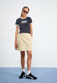 Tricou negru cu margini albe și logo-ul Puma, fustă înfoiată bej cu buzunare, adidași negri și șosete albe până la gleznă.