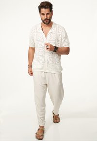 Weiße, kurzärmelige Strickbluse mit offener Struktur; lockere, hellbeige Hose; braune Sandalen; geschichtete Armbänder und Ringe als Accessoires.