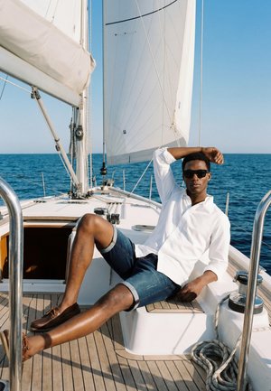 Homme portant des lunettes de soleil, une chemise blanche et un short en jean, se relaxant sur le pont d'un voilier avec l'océan et des voiles en arrière-plan.
