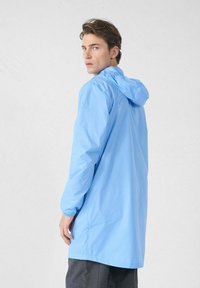 Junger Mann trägt eine hellblaue Kapuzenregenjacke und blickt über seine Schulter vor einem schlichten weißen Hintergrund.