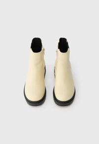 Béžové kotníkové boty s lesklým povrchem, kulatou špičkou, černou gumovou podrážkou a bočními zipy pro snadné obouvání. Hladký povrch s minimálními detaily.