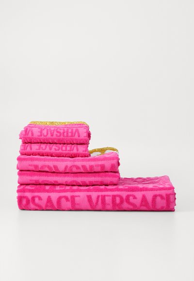 Une pile de serviettes roses vives avec le logo en relief "VERSACE" dans un motif texturé, avec une serviette supérieure accentuée d'or.