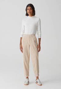 Witte langemouwtop, beige corduroy broek met elastische tailleband, twee zakken en cuffs bij de enkels. Gepaard met grijze en zwarte sneakers.